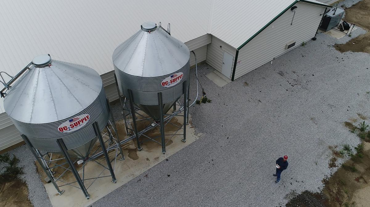A low altitude view of the exterior of a chicken barn. Feed bins and someone walking on the ground are visible.