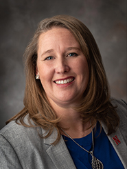 Smiling woman with long brown hair in a gray blazer and blue shirt against a neutral backdrop.