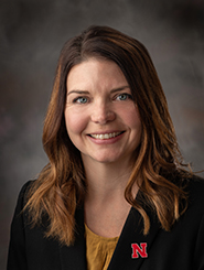 Smiling woman with shoulder-length brown hair wearing a black blazer and yellow shirt against a gray background.